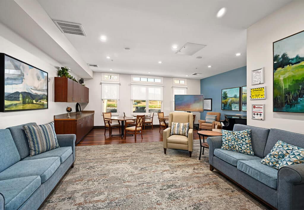 Comfortable common lounge area in a senior living building with sofas, chairs, and large windows.