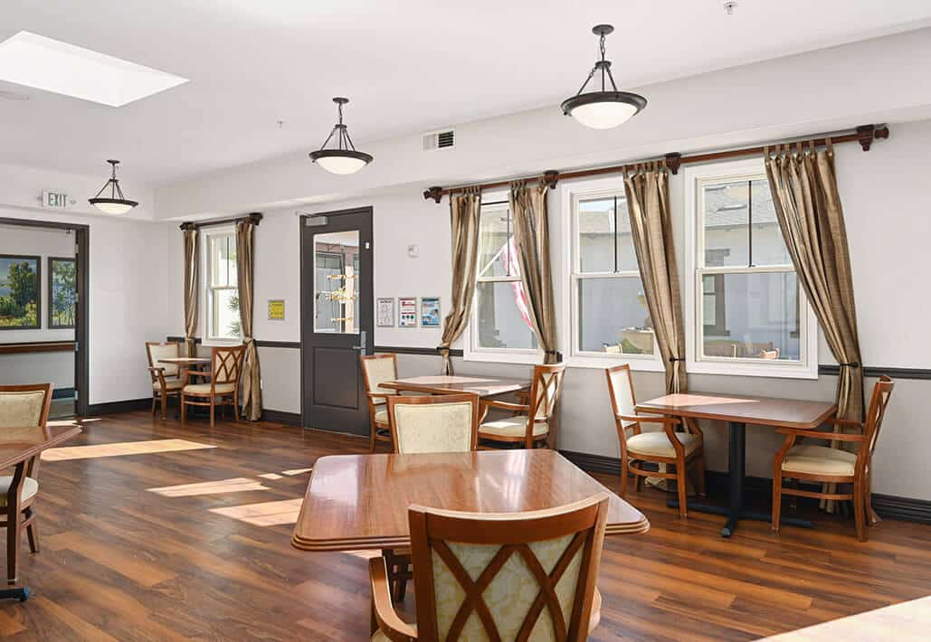 Sunlit communal dining area with wooden tables and chairs, and large windows with curtains.