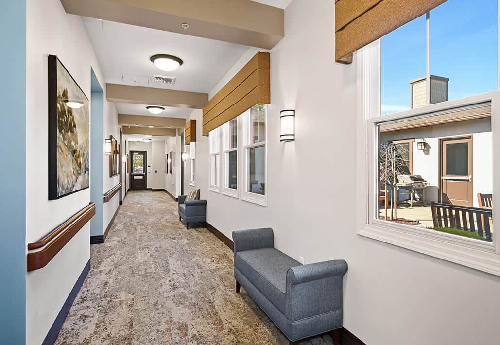 Bright hallway with seating and art, leading to outdoor courtyard at a senior living community.