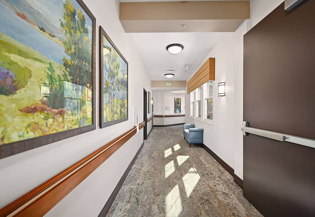 Well-lit hallway in a senior living community with landscape paintings and a blue armchair.