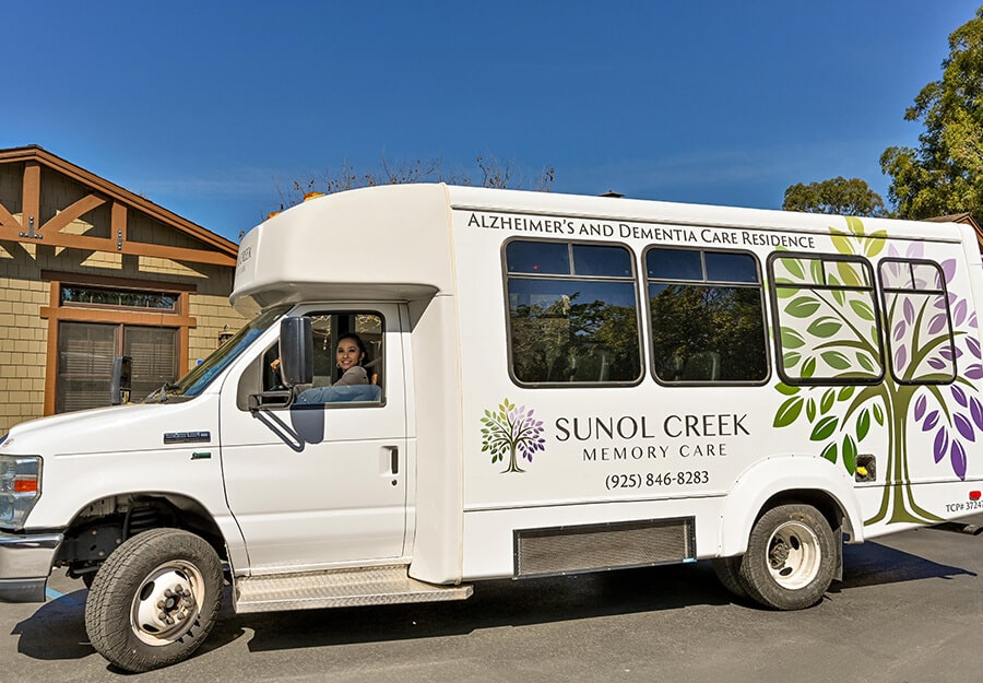 Memory care community shuttle with driver outside residential units on a sunny day.