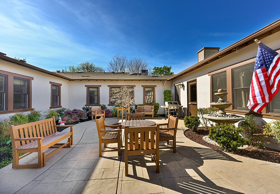 Outdoor courtyard with wooden benches, tables, and an American flag in a senior living community.